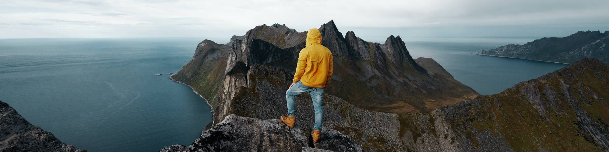 man in a yellow sweater stands on a cliff edge overlooking a montain range and sea